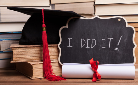 Little blackboard next to a diploma, some books, and a black mortarboard with red tassel.の写真素材
