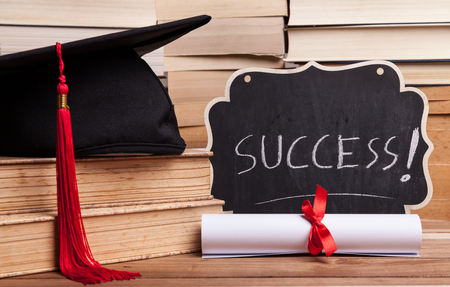 Little blackboard next to a diploma, some books, and a black mortarboard with red tassel.の写真素材