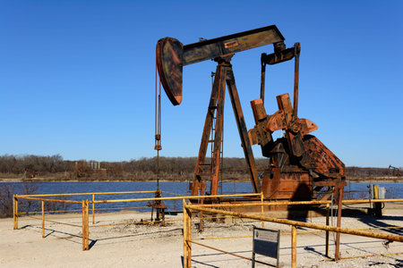 Rusty Oilfield Pumpjack (rocking horse) over a wellhead. Clear blue sky and lake background