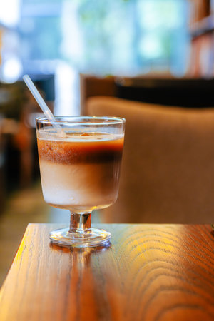 An iced latte in a glass sits on a wooden table in a warm-toned cafe filled with books, capturing a...