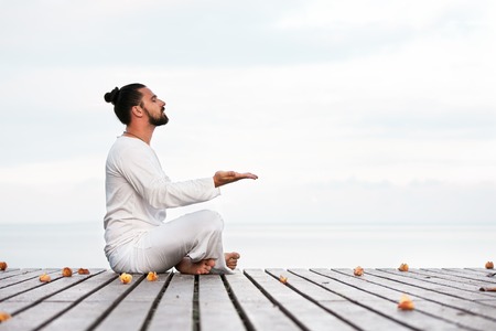 Caucasian man in white clothes meditating yoga on wooden pier platformの写真素材