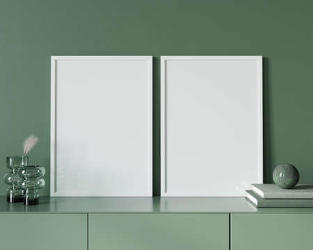 A poster with two white frames standing on a chest of drawers near a green glass vase with a wheat branch, books and a stone ball against the background of a green wall / 3D illustration, 3d renderの写真素材
