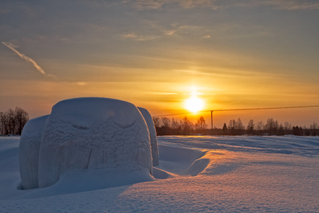The hay rolls on the fields of the Northern Finland have been covered by the snowfall. The winter sun sets beautifully in the background.の写真素材