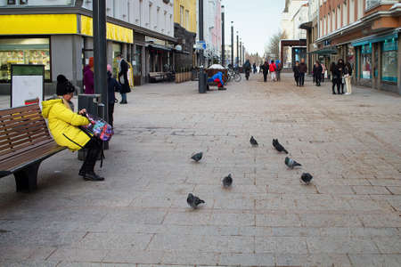 A woman in a yellow coat is feeding the pigeon on the street. She seems to be looking for more bird food in her bag.の写真素材