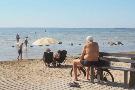 An older man enjoys a warm day at the beach. There are other seniors under a parasol and walking by.の写真素材