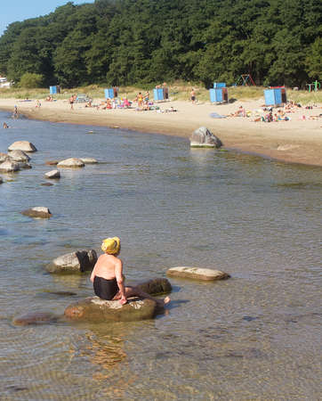 An elderly woman is sitting on a rock at the beach on a summer day. The warm day has lured a lot of people on the beach.の写真素材