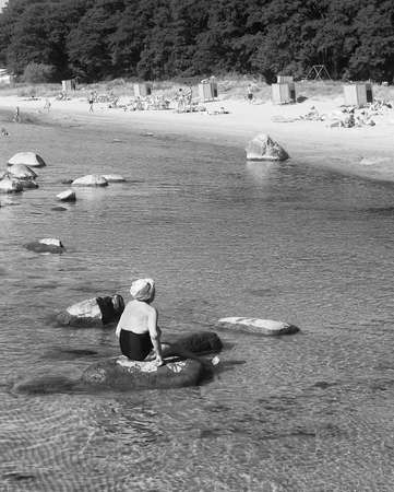An elderly woman is sitting on a rock at the beach on a summer day. The warm day has lured a lot of people on the beach.の写真素材