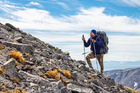European male tourist in sportswear with large backpacks and trekking poles climbs on stone scree to the top of Khibiny mountains. Group Hikers makes a sports hike in summer sunny day, Russia, Europeの写真素材
