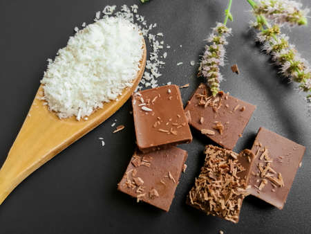 On a black background, pieces of chocolate, coconut chips and mint flowers