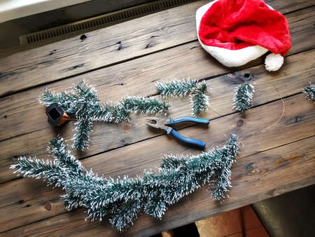 Making a Christmas garland with your own hands from artificial spruce branches. On the wooden table are twigs, pliers, wire and a Santa Claus hat.の写真素材