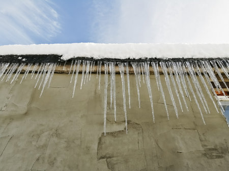 Dangerous sharp icicles hang from the roof. Beautiful icicles made of melted snow. Transparent large icicles on the edge of the roof, it is dangerous to walk.の写真素材