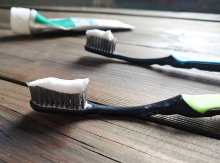 Two toothbrushes with toothpaste on them lie on a wooden table. In the background is a tube of...
