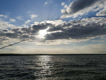 the abandoned lure merges with the sun. Viid on the river. fishing on the river - the fishing rod is thrown into the water. A fishing rod and a river with cloudy skies.の写真素材