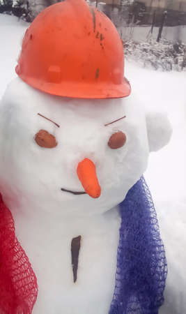 A snowman in an orange helmet with walnut shell eyes and a mesh scarf. an orange carrot like the nose of a snowman. Gloomy snowman. An evil snowman with an ear.の写真素材