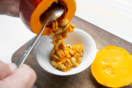 A person cleans a pumpkin from seeds. Pumpkin peeling. Pull the seeds from the pumpkin with a metal spoon into a plate. Cooking pumpkin.の写真素材