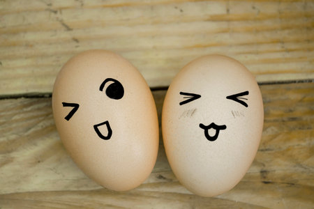 Two eggs with emoticons on the background of a wooden board. On a wooden table are two eggs with emoji. On the eggs, the drawings are emotions. A smile and a wink at the eggs.の写真素材