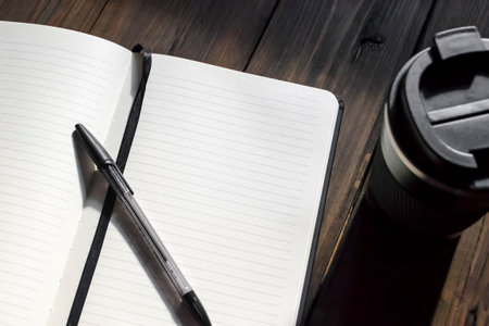 Notebook with a pen on a wooden table next to a cup of coffee. Writing utensils and coffee making facilities. Open notepad with pen.の写真素材