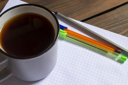 A cup of coffee next to the notepad and pens. On the table is a plaid notebook, three pens and a cup of black coffee. Americano during his studies.の写真素材