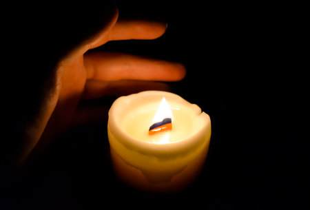 In the dark, a hand reaches for a flame candle. The warmth of the candle warms the skin. The faint light of the candle illuminates the hand. Hand and candle in a dark room.の写真素材