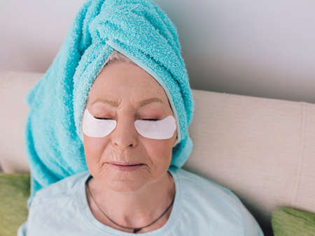 mature caucasian stylish woman laying on a coach with blue towel on her had and collagen eye patches. Anti age, healthy life style,の写真素材