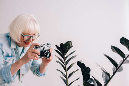 senior (old) stylish woman with gray hair and in glasses and jeans jacket taking pictures of flowers with film camera. Age, hobby, anti age, positive vibes, photography conceptの写真素材