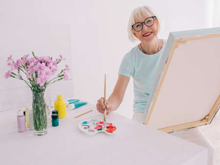 senior cheerful woman in glasses with gray hair painting still life. Creativity, art, hobby,...