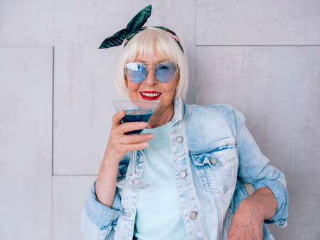 senior (old) stylish woman with gray hair and in blue glasses and denim jacket holding glass with blue cocktail. Alcohol, relax, holidays, retirement conceptの写真素材