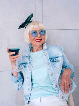 senior (old) stylish woman with gray hair and in blue glasses and denim jacket holding glass with blue cocktail. Alcohol, relax, holidays, retirement conceptの写真素材