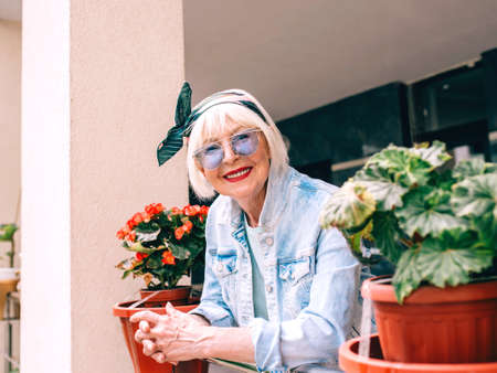senior (old) stylish woman with gray hair and in blue glasses and denim standing on balcony...