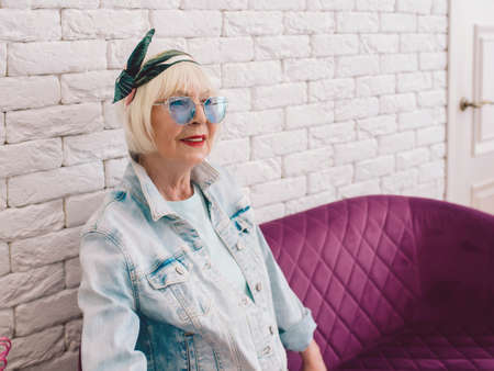 elderly senior stylish woman in blue sunglasses and jeans jacket sitting on the sofa in stylish loft...
