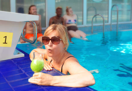 funny emotional blonde woman by the edge in the swimming pool with green cocktail and with friends....