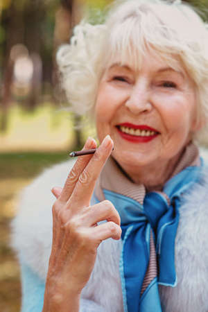 smiling senior elegant stylish fashionable woman with grey hair in fur coat outdoor smoking cigarette. Unhealthy lifestyle, age, positive vibes, addiction, bad habit concept.の写真素材