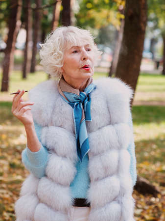 sad fashionable senior woman with grey hair in fur coat outdoor smoking cigarette. Unhealthy lifestyle, age, oldness, addiction, bad habit concept.の写真素材