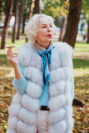 sad fashionable senior woman with grey hair in fur coat outdoor smoking cigarette. Unhealthy lifestyle, age, oldness, addiction, bad habit concept.の写真素材