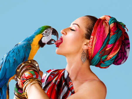 Portrait of young attractive woman in african style holding ara parrot on her hand and kissing it by...