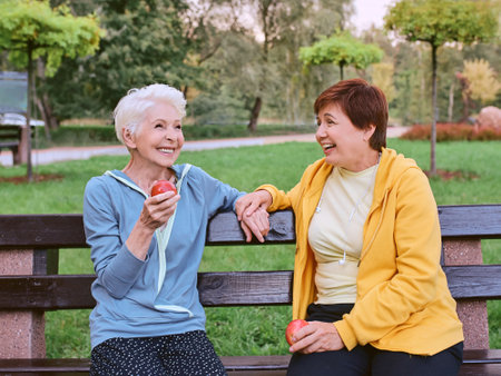 two mature women eating apples on the bench after doing sport exercises in the park. healthy lifestyle conceptの写真素材