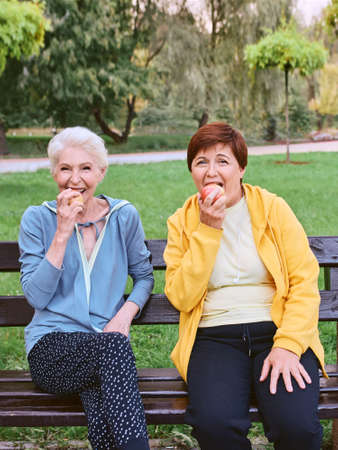 two mature women eating apples on the bench after doing sport exercises in the park. healthy...