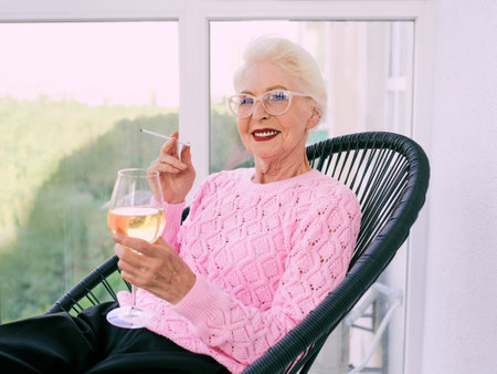 Old fashioned senior stylish woman sitting on terrace smoking cigarette with glass of white wine. Bad habit, addiction conceptの写真素材