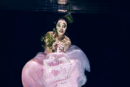 young beautiful woman in long pink dress with cage in her hands underwaterの写真素材
