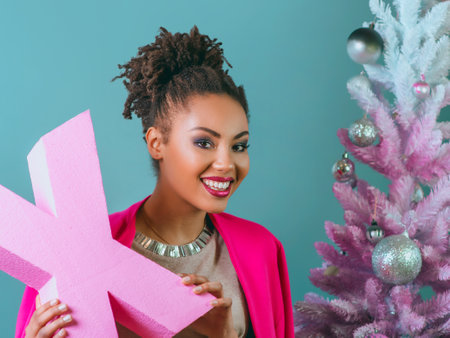 cheerful afro american woman with pink letter X in her hands on the christmas tree background. Christmas, new year, happiness, holidays conceptの写真素材