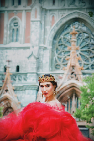 Beautiful young woman in long red dress and in royal crown nearly catholic cathedralの写真素材