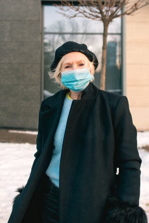 senior stylish woman in beret and elegant black coat and in medical mask walking outdoor. Lockdown,...