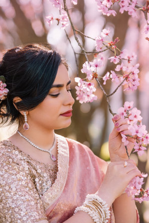A woman in a shimmering pink traditional outfit stands serenely among cherry blossoms, radiating warmth, elegance, and cultural pride.の写真素材