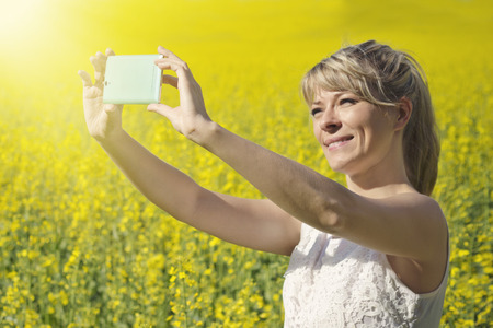 Technology, summer holidays, vacation and people concept - hands of woman taking selfie by smartphone on rape fieldの写真素材