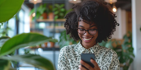 Joyful black woman using phone to communicate or social media in officeの素材