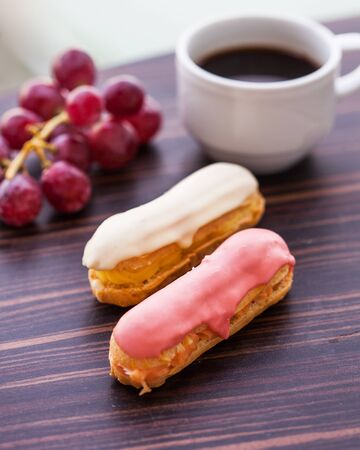 Delicious eclairs close-up with pink and white glaze, in the background a white mug with black coffee and a bunch of grapes.の写真素材