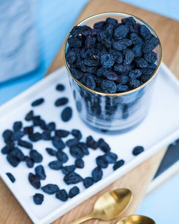 Delicious black raisins in a transparent glass on a white tray and a blue background. Healthy snack.の写真素材