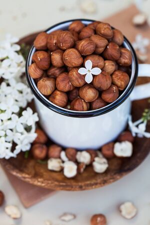  A handful of peeled hazelnuts in a white mug on a wooden plate, next to a white lilac flowers. Food photo of a healthy snack, veggie snacks.の写真素材