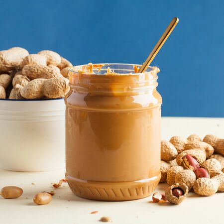 An open jar of peanut butter with a spoon and peanuts in the shell around on a colored background....