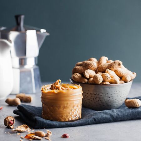 Closeup peanut butter in a glass jar, a handful of peanuts in shell on a gray background. Foodphoto....
